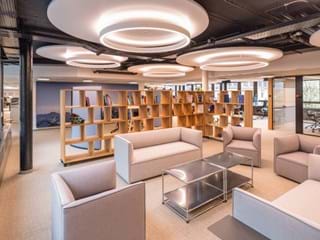 Modern office space featuring large circular acoustic ceiling panels with lights, beige sofas arranged around a metal coffee table, and wooden bookshelves with various books. Work stations and a meeting room are visible in the background. Large windows allow natural light to illuminate the room, creating an inviting and open atmosphere.