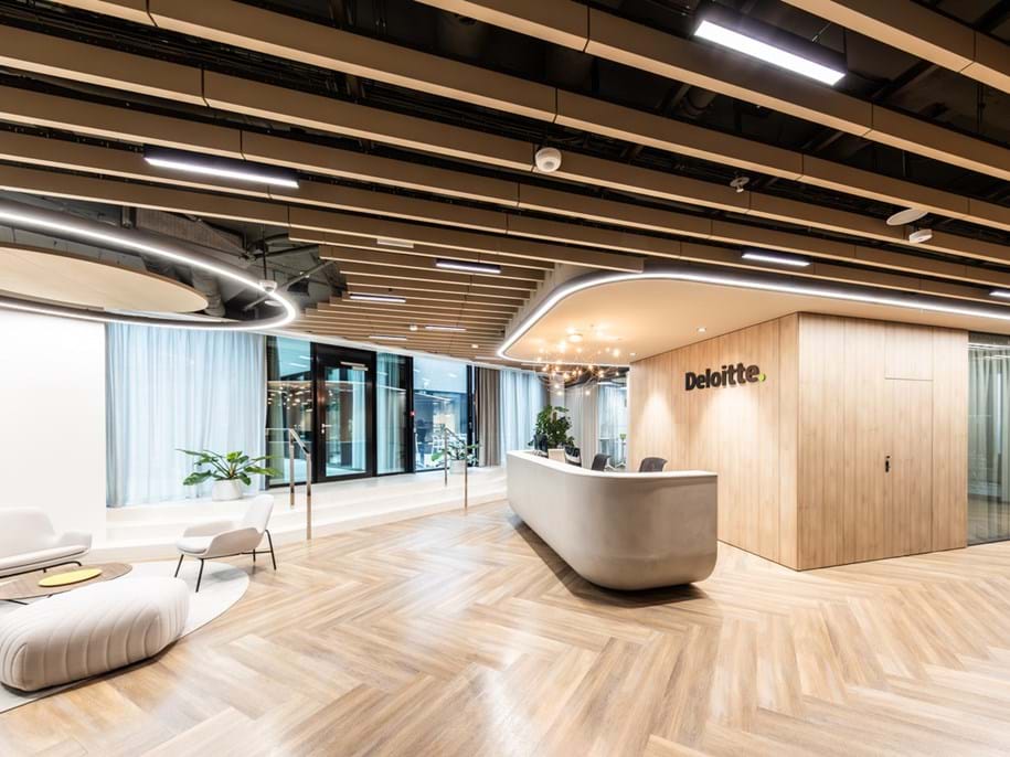 A modern office lobby with the Deloitte logo on the wall behind a curved reception desk. The floor is covered with light wooden planks, and the ceiling features hanging light-brown acoustic baffles and curved lighting fixtures. To the left, there is a seating area with a couch and two chairs around a low table. Large windows allow natural light to fill the space, creating a welcoming atmosphere.