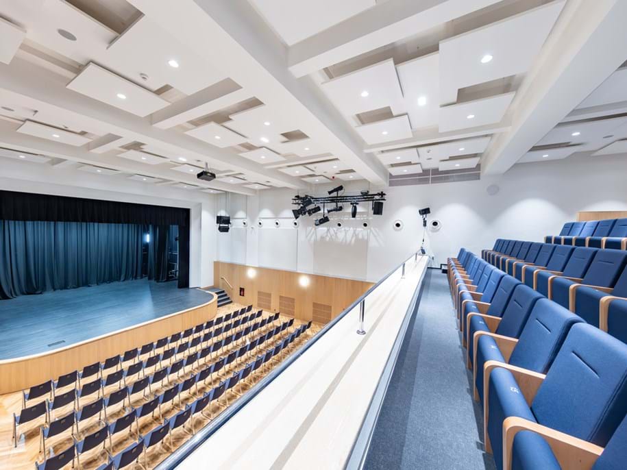 An empty auditorium with a modern design, featuring rows of blue upholstered seats, wooden panels on the walls, and a stage with a closed blue curtain. The ceiling has geometric acoustic panels with integrated lighting, and there are multiple speakers mounted near the ceiling. The perspective is from the back of the auditorium looking towards the stage.