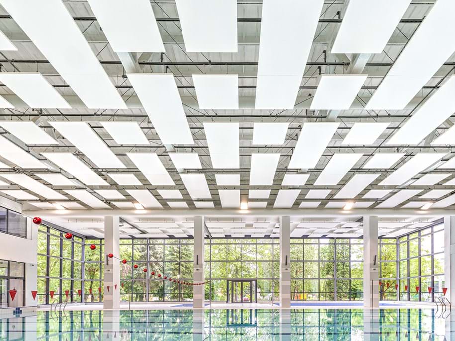 Interior of a modern building featuring a large swimming pool. The ceiling is composed of an intricate geometric pattern with numerous square and rectangular acoustic panels. Natural light floods the space through floor-to-ceiling windows, which are evenly spaced along the walls. Red spherical flags hang at intervals across the pool, adding a pop of colour to the predominantly white and grey tones of the room.