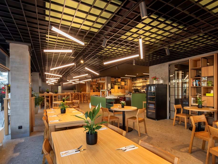 Modern restaurant interior with wooden tables and chairs, minimalistic decor, and plants. Bright lighting fixtures hang from the grid-patterned ceiling. The background features a bar area and shelving with various items.