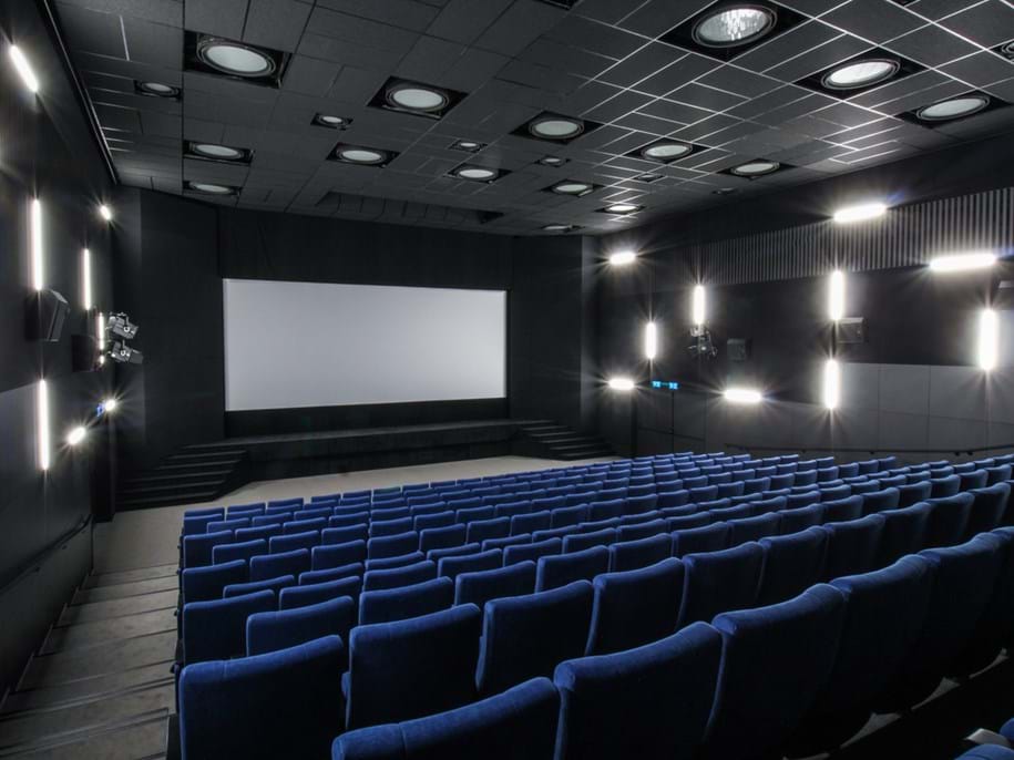 Modern movie theatre with rows of blue seats facing a large blank screen. The walls and ceiling are dark, equipped with circular lights and speakers. The room is empty, creating a quiet and spacious atmosphere.