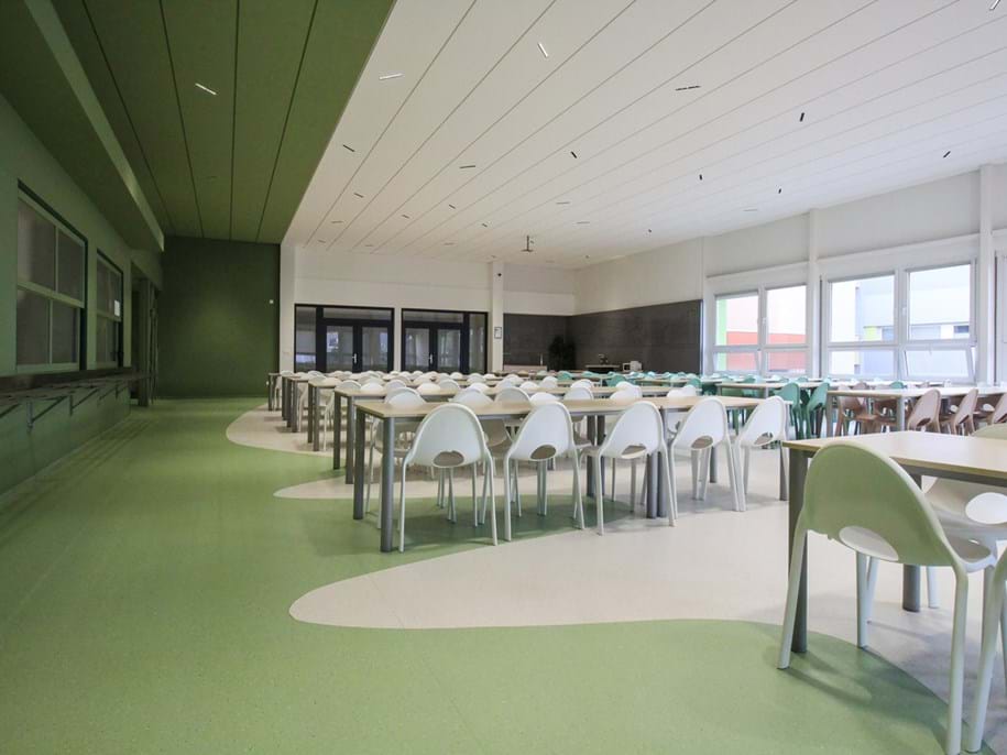 A spacious, modern canteen with rows of white tables and chairs, large windows letting in natural light, and green and white flooring and walls. The room is empty and neatly arranged.