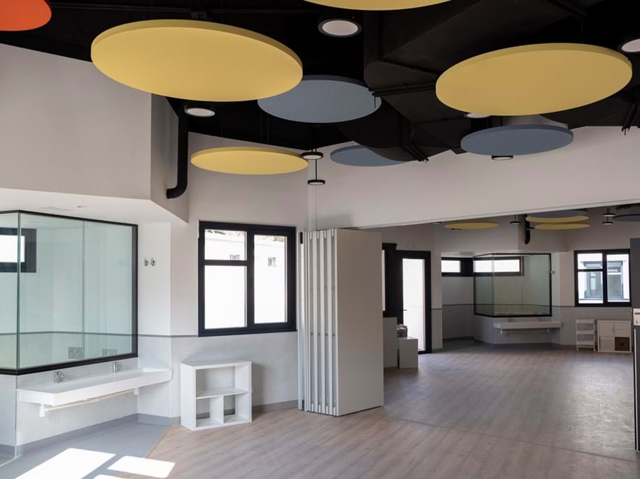 A modern, spacious room with wooden flooring, bright overhead lighting, and circular acoustic panels in yellow and blue on the ceiling. The room features a sink, large windows, and white walls with a partition for dividing spaces.