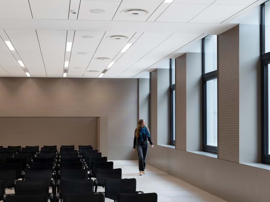 White, suspended acoustic ceiling in large conference room with beige floor and walls, black chairs in multiple rows and a woman looking out the big windows
