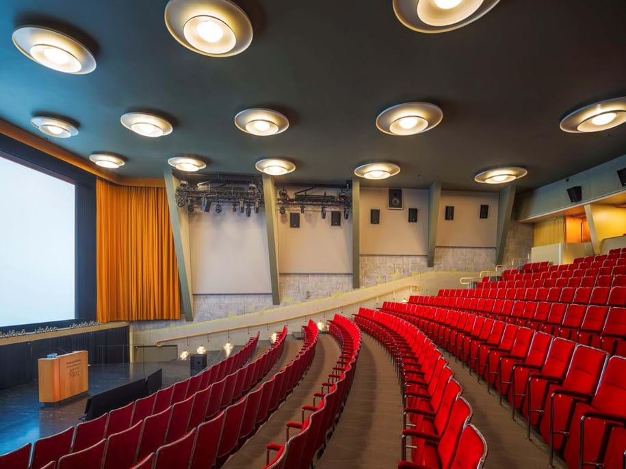 Acoustic stretch fabric wall panels in classic cinema with rows of chairs in red fabric and a petrol blue ceiling with round lamps