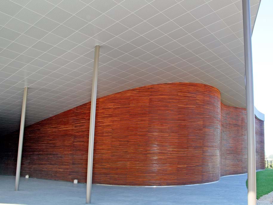 Curved wooden wall with slatted design beneath a modern, grid-patterned metal ceiling. Three metal columns support the structure, with a concrete floor and a glimpse of greenery outside. The design is sleek and contemporary.