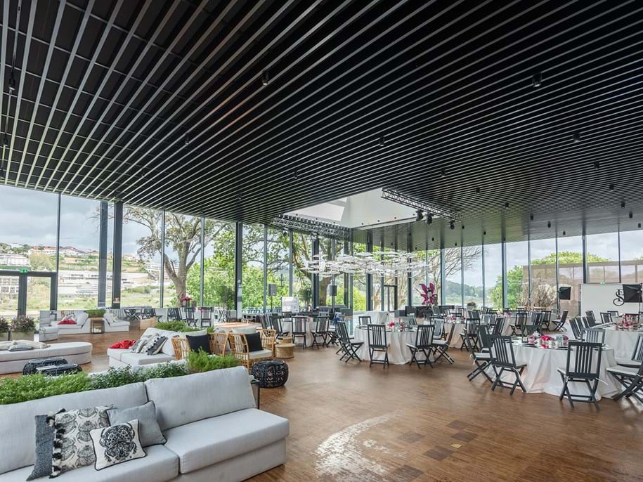 Spacious modern event venue with large windows, wooden floor, and black ceiling slats. Round tables are set with white linens and black chairs. Couches with pillows are arranged by the windows. Scenic views of greenery and hills are visible outside.