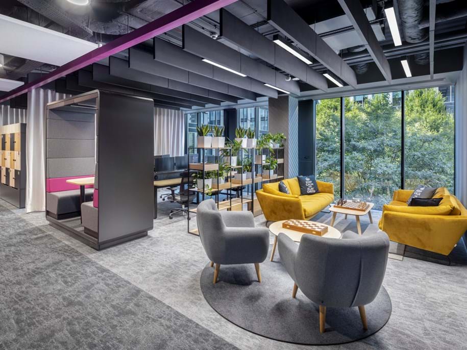 Modern office space with grey and mustard yellow seating, small round tables, and potted plants. Floor-to-ceiling windows offer a view of greenery outside. The ceiling features exposed ducts and purple accents. Open workstations are visible in the background.