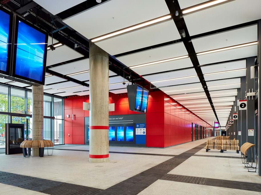 Modern bus station interior with high ceilings, red and gray walls, digital information screens, and wooden benches. Bright lighting illuminates the space, with a long corridor visible in the background. Signage with numbers hangs from the ceiling. Large entrance doors are visible to the left bringing daylight into to the terminal.