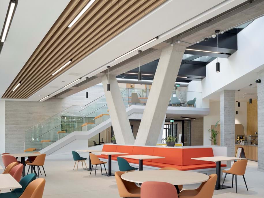 Modern interior space with high ceilings and natural light, featuring tables and colourful chairs. Acoustic ceiling baffles complement the V-shaped concrete pillars, and a staircase with glass railings to enhance the contemporary design.