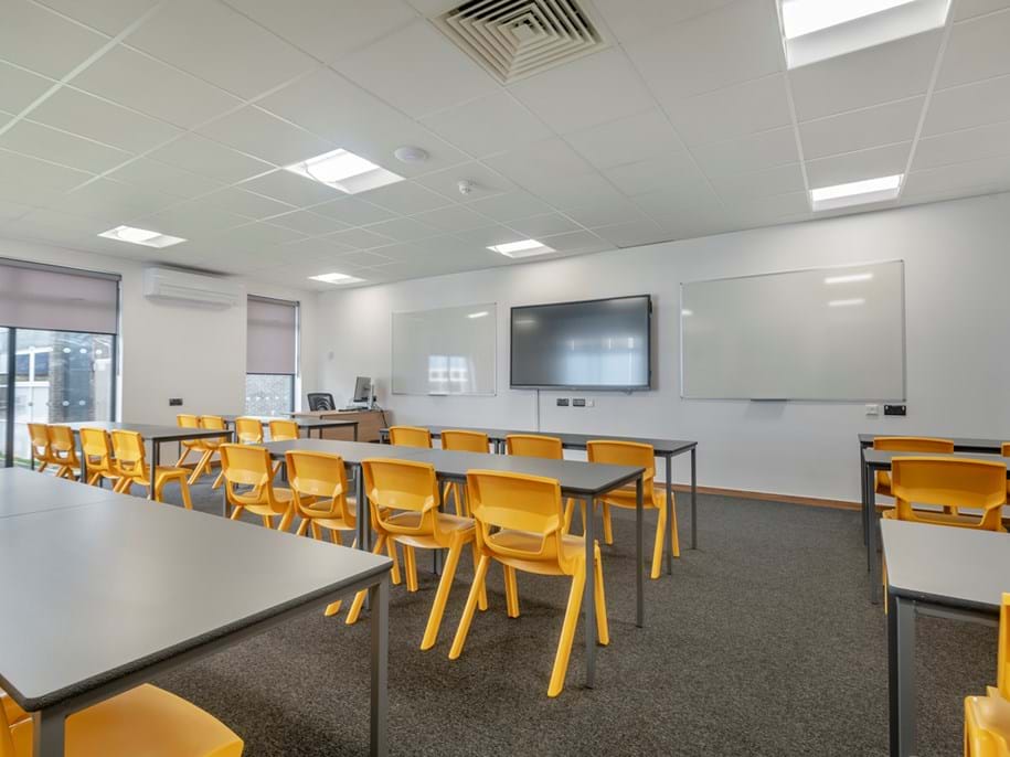 A modern classroom with a whiteboard and digital screen on the wall, multiple rows of yellow chairs and desks facing forward, and large windows allowing natural light to fill the room. The white acoustic ceiling features integrated lighting. The setting appears to be ready for an educational session or lecture.
