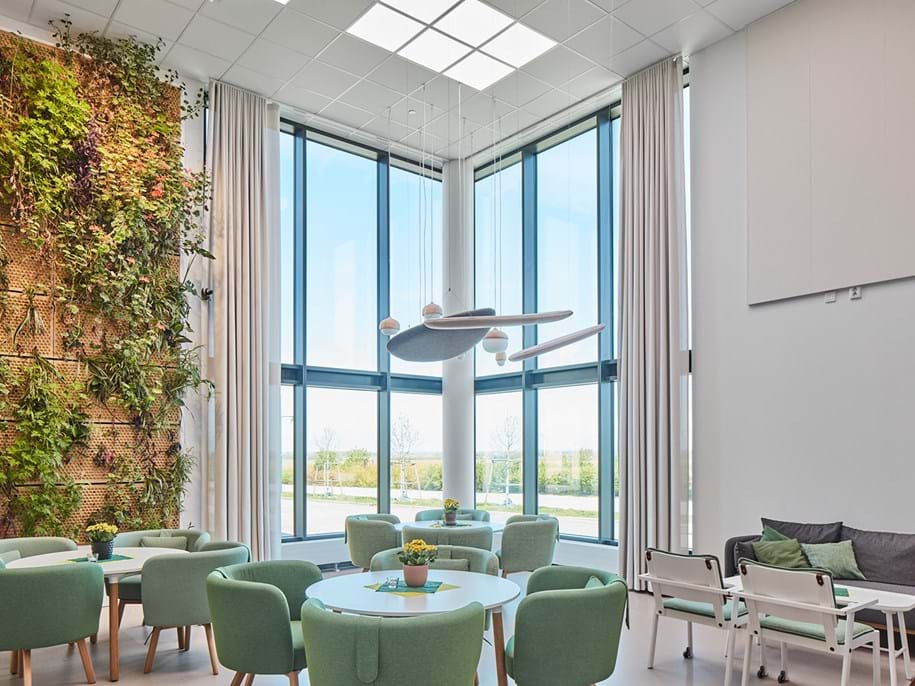A modern and bright room with large windows providing ample natural light. The room features a living green wall with various plants on the left side, adding a touch of nature indoors. In the center, there are round tables surrounded by mint green chairs with a yellow floral centerpiece. Above the table hangs an artistic light fixture with multiple round lights at different heights. To the right, there is a cozy seating area with grey sofas and chairs around a low coffee table. The overall atmosphere is clean, refreshing, and designed for relaxation or casual meetings.