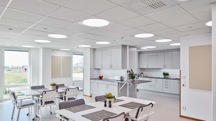 Una cucina moderna e ben illuminata con pareti e soffitto bianchi. Lo spazio presenta diversi tavoli bianchi con sedie, alcuni decorati con tovagliette grigie. L'area della cucina include mobili bianchi, elettrodomestici in acciaio inossidabile e un'isola centrale con lavello. Piccole piante in vaso aggiungono un tocco di verde, e ampie finestre permettono l'ingresso di molta luce naturale. La stanza è illuminata da luci a incasso nel soffitto, creando un'atmosfera pulita e accogliente.