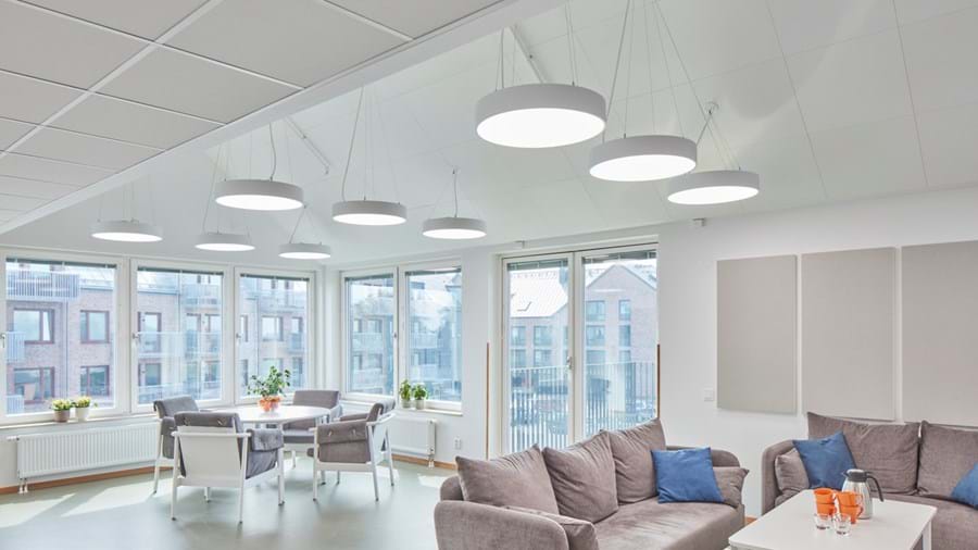 Una moderna sala relax ben illuminata con grandi finestre che offrono una vista sull'esterno. La stanza è arredata con un comodo divano grigio con cuscini blu, diversi tavoli bianchi abbinati a sedie e lampade circolari a sospensione. Le pareti sono rivestite con pannelli acustici bianchi, creando un'estetica pulita e minimalista, ideale per il relax.