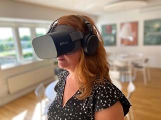 Žena s dlouhými kaštanovými vlasy má na sobě náhlavní soupravu pro virtuální realitu Pico a sluchátka. Je ve světlém, moderním pokoji s dřevěnými podlahami a bílým nábytkem. Na stěnách na pozadí se zobrazí rozmazaná umělecká díla.