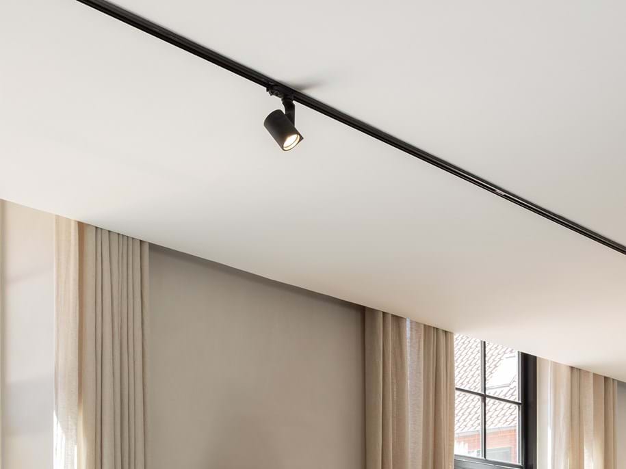 Close-up of a white, smooth ceiling with a black lighting track fitted to it. Two windows with beige curtains are visible in the background.