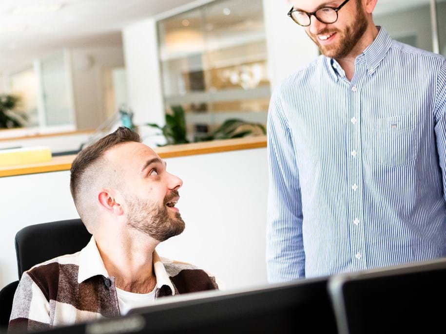 Twee mannen met baarden bevinden zich in een kantooromgeving. De een zit aan een bureau en lacht naar de ander, die staat en een bril draagt. Ze lijken een vriendelijk gesprek te voeren. Er zijn verschillende computerschermen en kantoordecoraties zichtbaar.