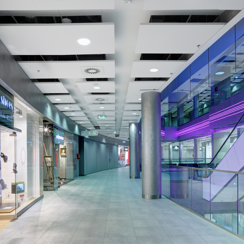 Modern shopping mall corridor with glass storefronts on the left and a blue-lit escalator on the right. The floor is tiled, and the ceiling features recessed lighting. The scene is brightly lit and spacious.