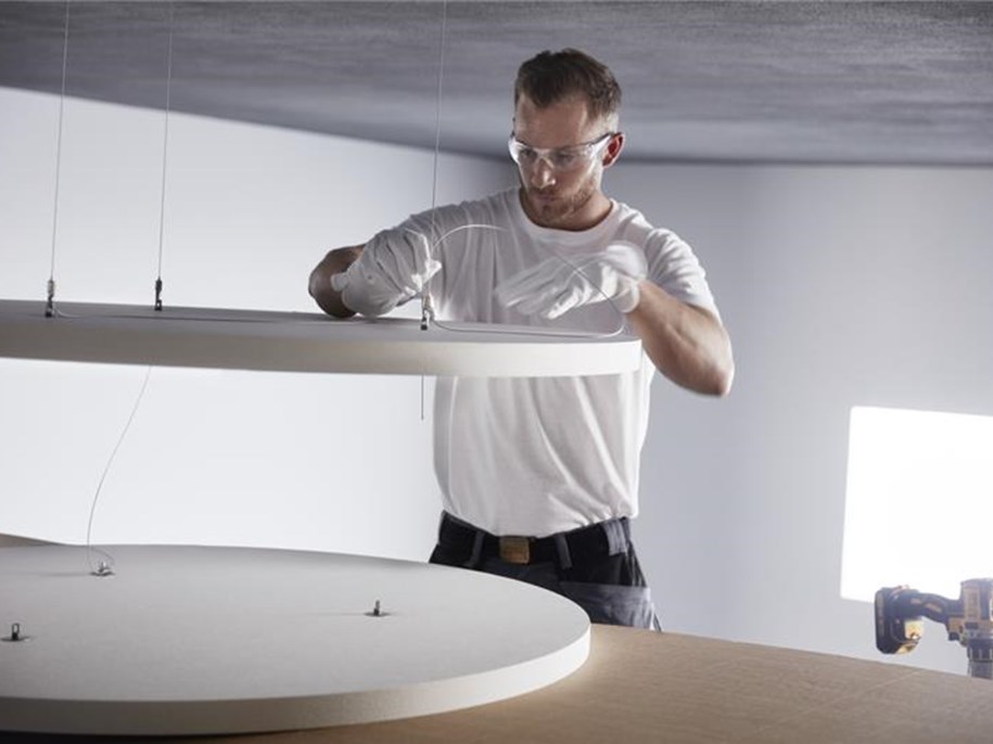 A man wearing safety goggles and gloves is adjusting a large circular acoustic panel suspended by wires in a bright room. A drill rests on the wooden surface nearby.