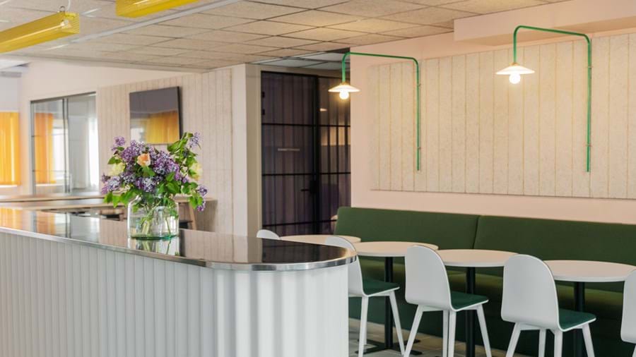 Reception area with suspended acoustic wood wool ceiling, yellow and green lamps, white and green furniture and a vase with flowers on the reception desk