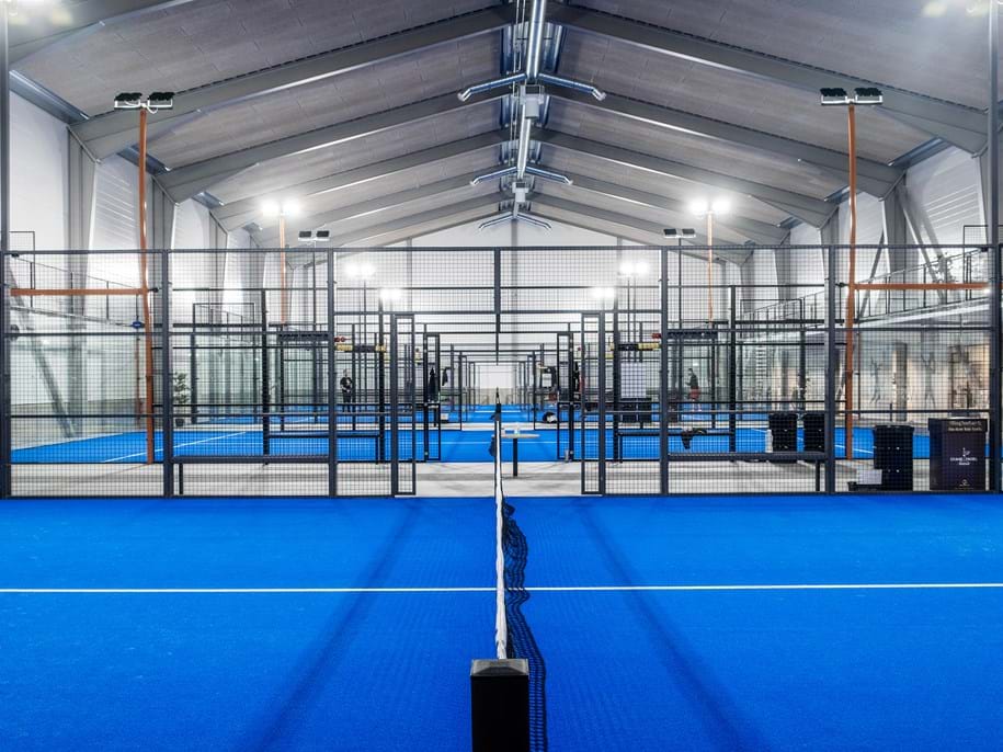 A large indoor padel court with bright blue flooring and a high, vaulted ceiling. The image shows multiple courts lined up side by side, surrounded by glass walls and metal nets, illuminated by overhead lights.