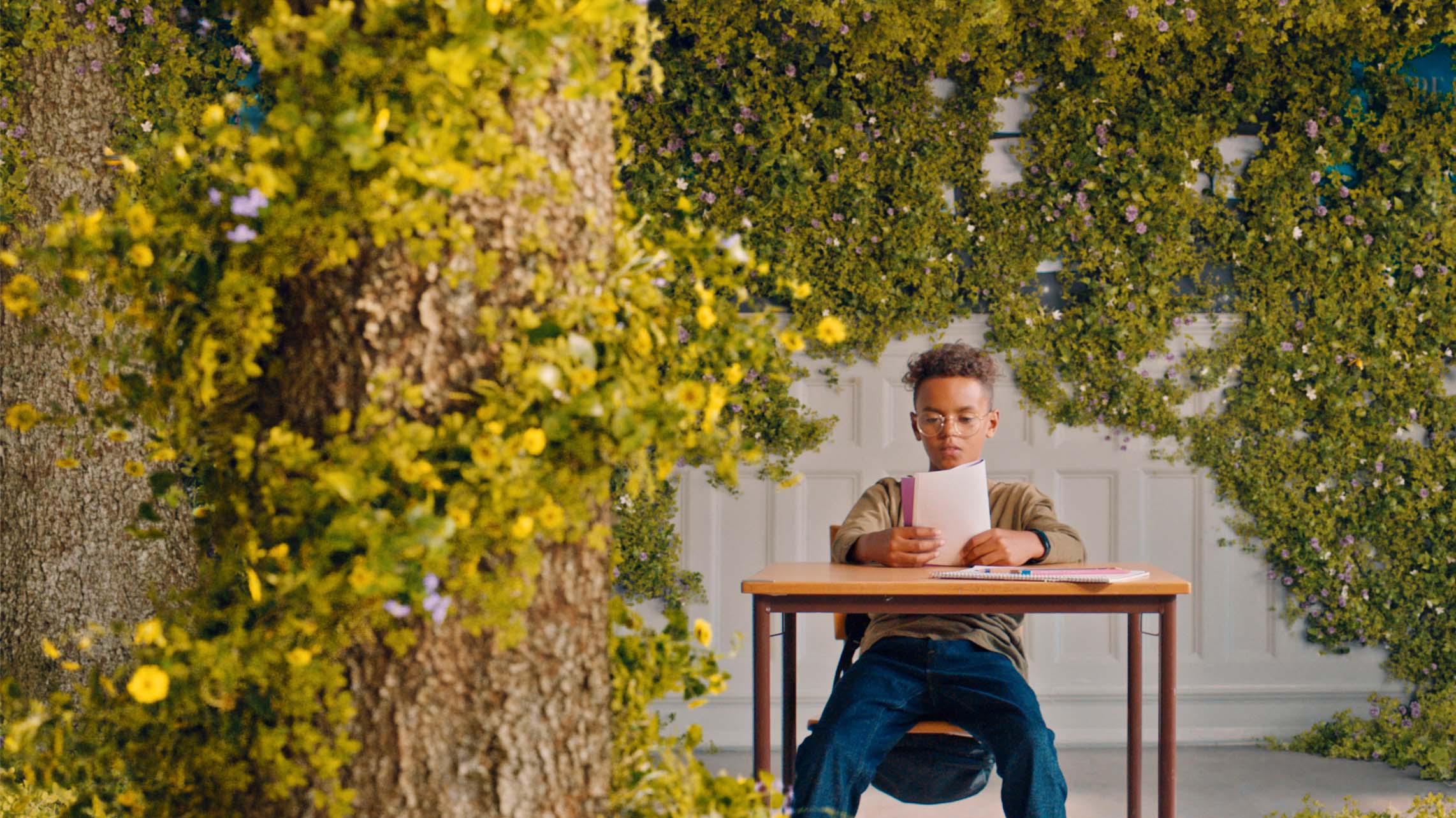Boy reading a book at school desk in room with plants growing on the walls