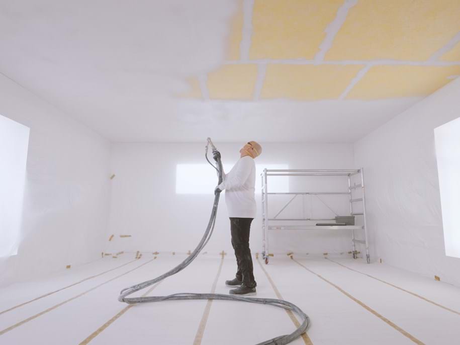 A person in a white room spraying acoustic plaster to the ceiling. The room has large windows, and there is a metal scaffold on wheels in the far right corner. The person is wearing protective clothing and a mask, holding a hose that is connected to the spray equipment. Yellow acoustic boards are visible on parts of the ceiling, contrasting with the white walls and floor that are covered with protective plastic sheeting.