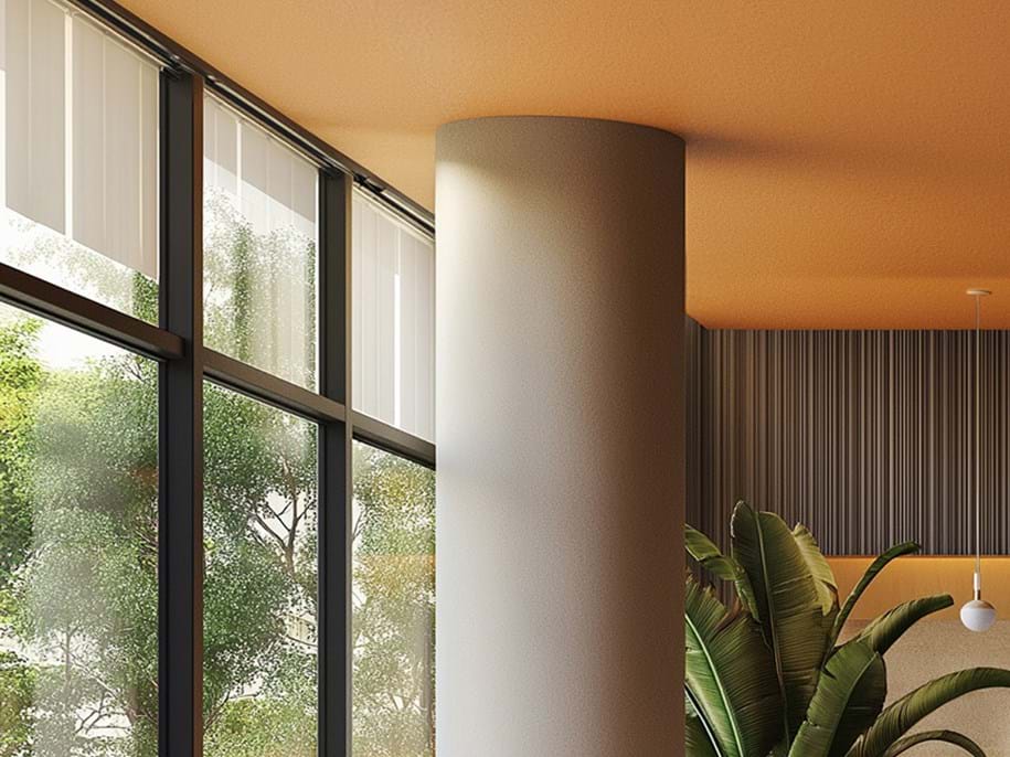 Interior view of a modern room with large windows revealing green trees outside, a cylindrical column in the centre, and a potted plant to the right. The room has an orange ceiling, white pendant light, and vertical blinds.