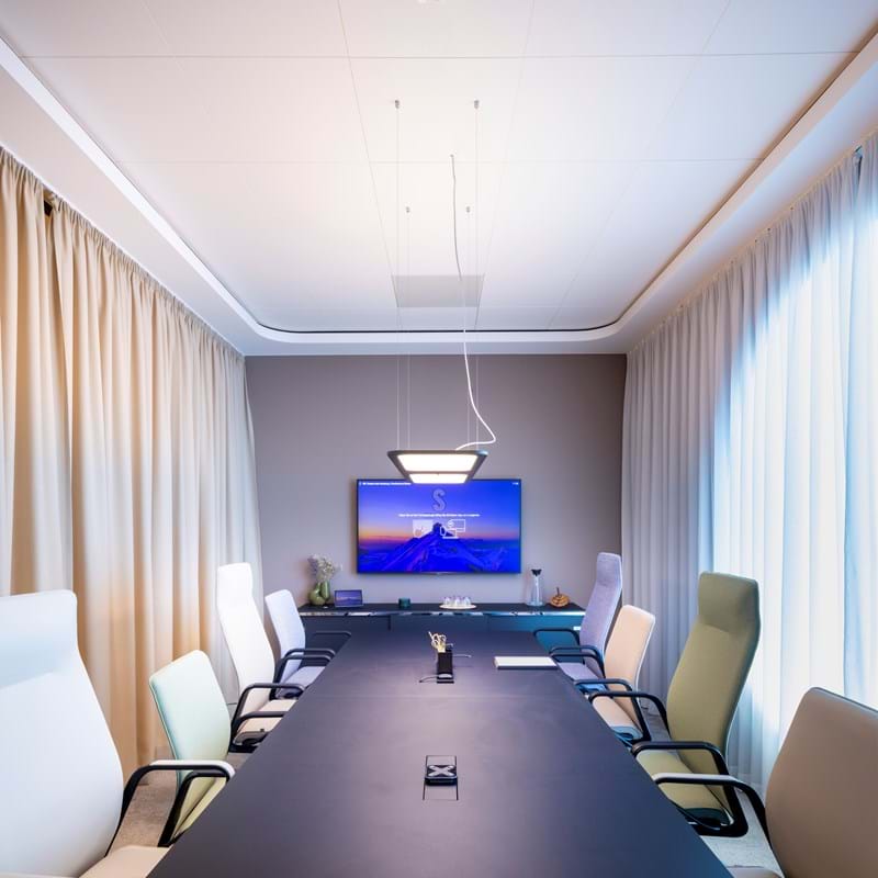 White, suspended acoustic ceiling in large conferene room with chairs in light colours, curtains covering the windows and glasswalls and a screen on the wall