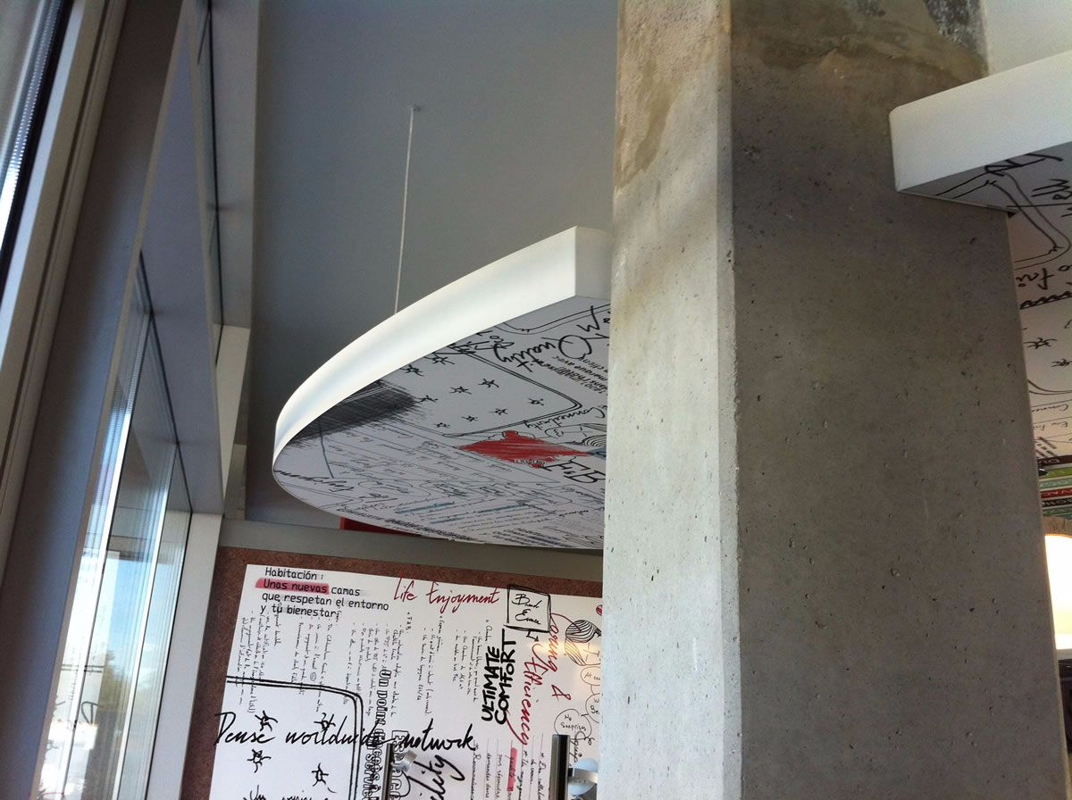 A modern interior features a large concrete pillar and a partial view of a circular ceiling panel covered in various scripts, doodles, and symbols. Natural light streams in from tall windows on the left. The style is contemporary and artistic.
