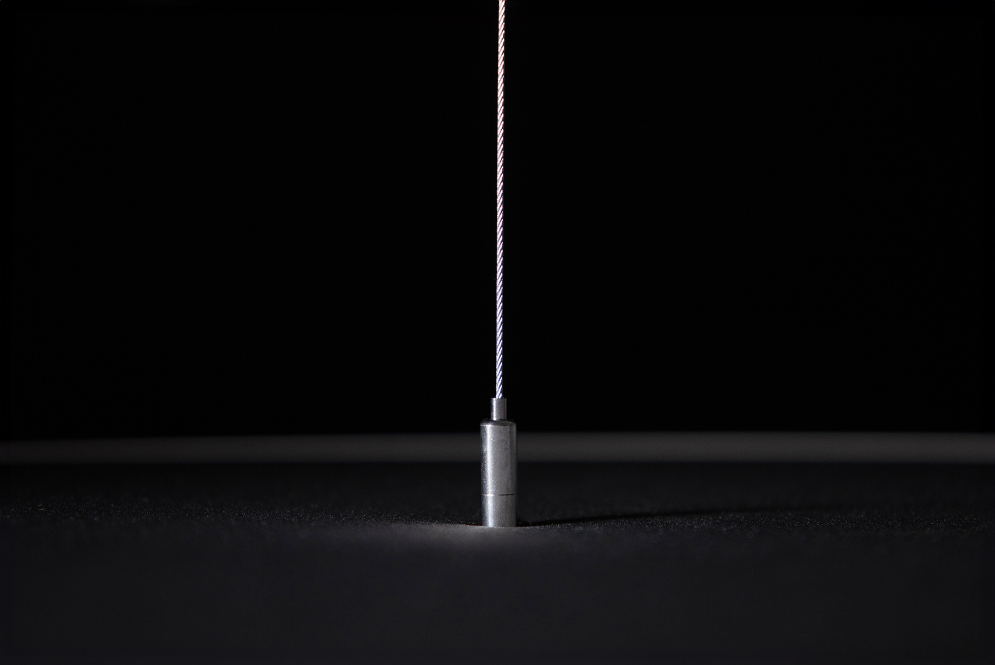 A glowing thin wire descends from above into the center of a metallic cylinder set against a dark background. The minimal lighting emphasises the texture and sheen of the materials.