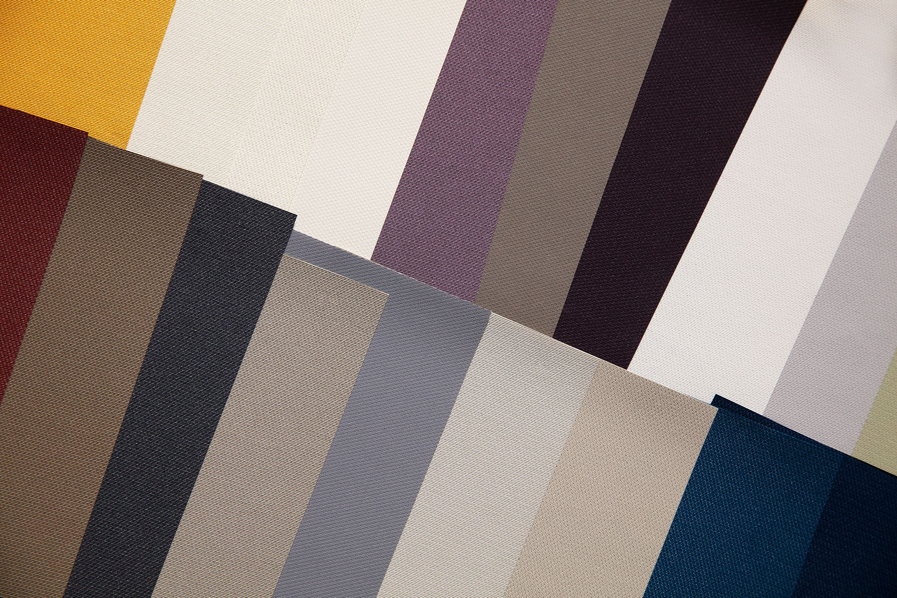  A collection of fabric samples displayed at angles, featuring a variety of colours including yellow, grey, beige, blue, purple, pink, and brown. The textures vary subtly, showcasing different fabric weaves.