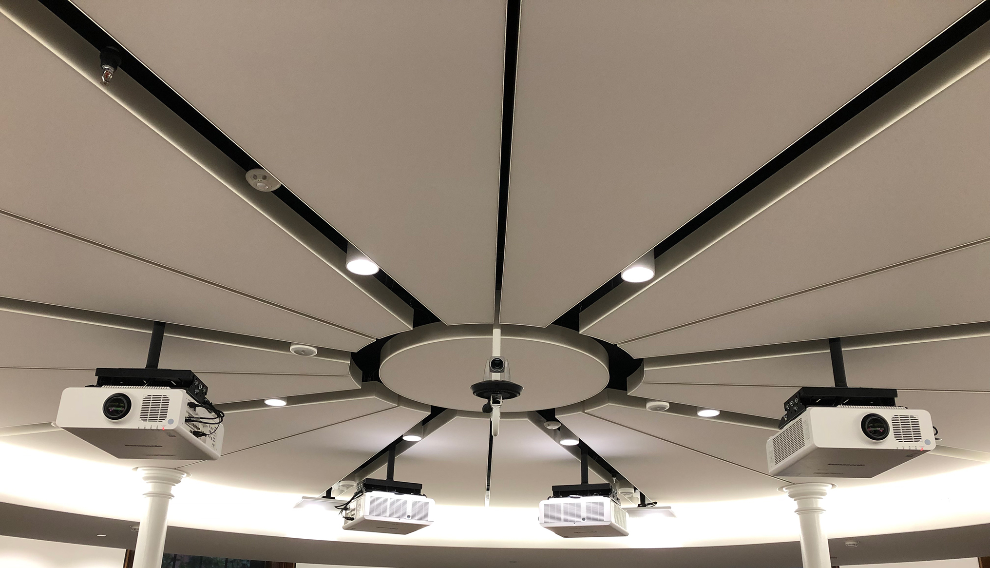Ceiling view of a circular conference room with a central camera surrounded by several projectors mounted on beams radiating outward, resembling spokes of a wheel. The ceiling is light coloured with recessed and spotlighting.
