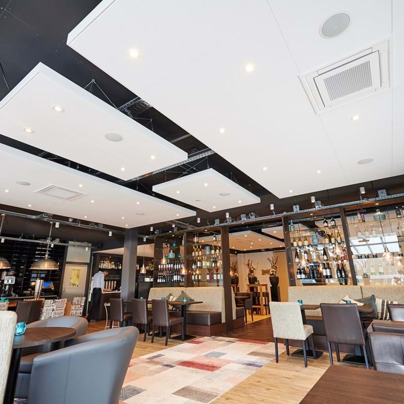 A modern restaurant interior with a mix of brown and beige chairs around dark wooden tables. The ceiling features large white panels and hanging round lights. A wall-mounted wine rack is visible in the background.