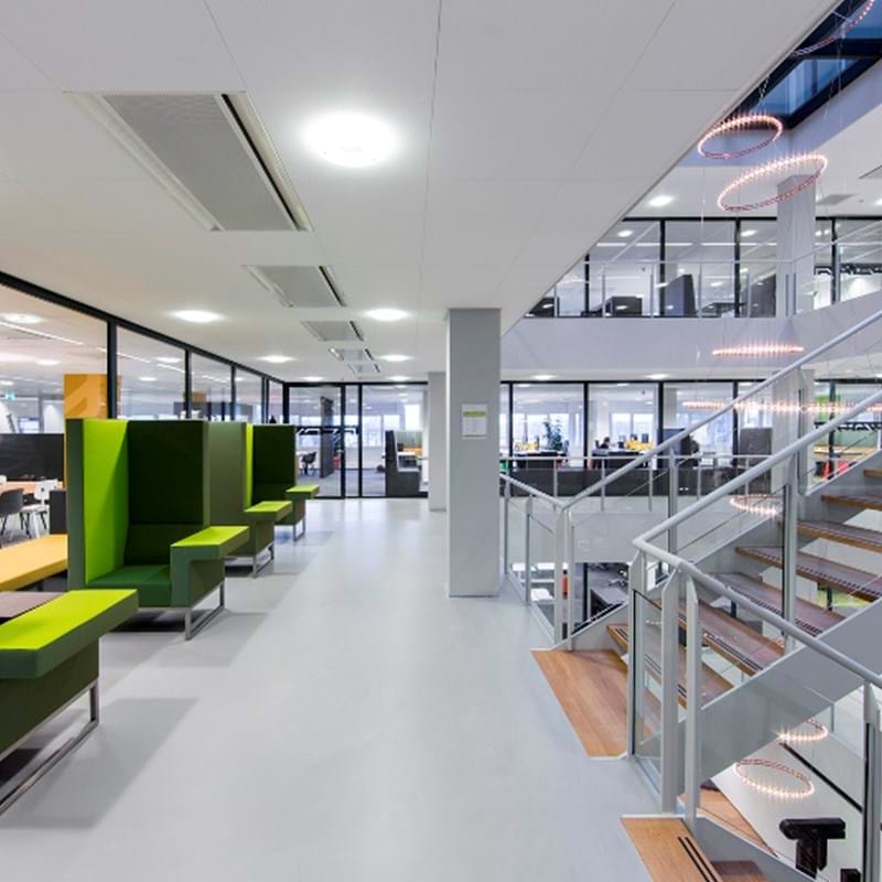 Modern office interior with an open layout. Green seating booths line the left side, while a glass-walled staircase on the right leads to an upstairs area. Natural light fills the space through large windows.