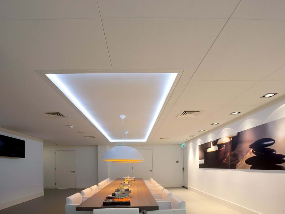 A modern conference room with a long wooden table and white chairs. A large pendant light hangs above the table. The room features recessed ceiling lights and a large wall art piece with abstract designs. A wall-mounted TV is on the left.