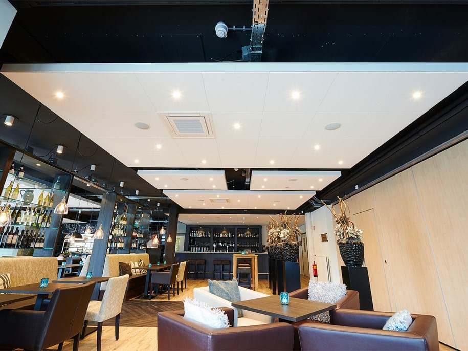 Modern restaurant interior with tables and cushioned chairs, featuring ambient lighting and a well-stocked bar in the background. The ceiling has recessed lights and decor includes vases with dried plants. The atmosphere is cozy and inviting.