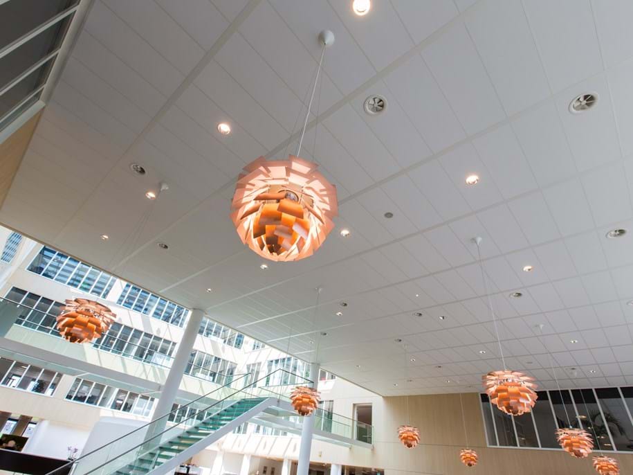 Spacious atrium with modern design features, including multiple copper pendant lights hanging from the ceiling. Large windows provide natural light, and a glass staircase leads to an upper level. The area has a sleek and contemporary aesthetic.