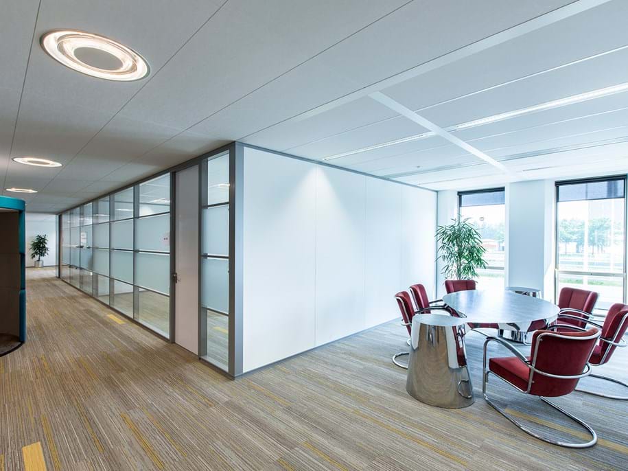 A modern office space featuring a round conference table with red chairs, adjacent to large windows. The room has grey and white walls, a wooden floor, and potted plants. Frosted glass panels separate the spaces, and ceiling lights illuminate the area.