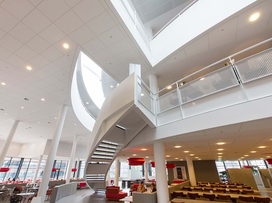 Spacious modern interior with multi-level open design, featuring a prominent white staircase. Large windows and a high ceiling enhance brightness. Red-accented seating is below, creating a sleek and welcoming atmosphere.