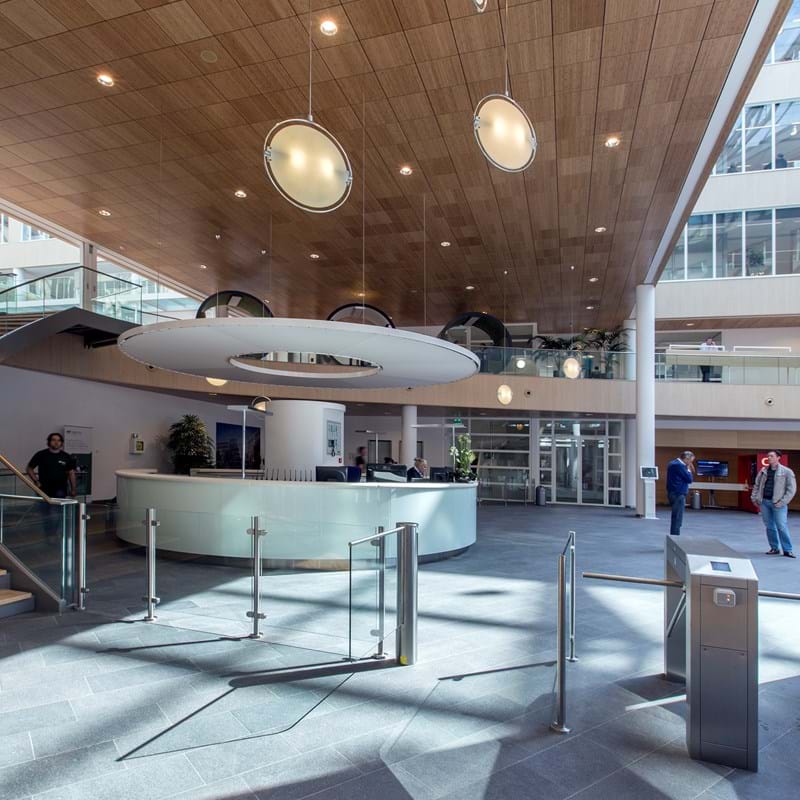 Spacious lobby with modern design, featuring a wooden ceiling with circular lights, a curved reception desk, and glass entrance doors. Floor is grey tile, and several people are standing or walking. Metal turnstiles are near the entrance.