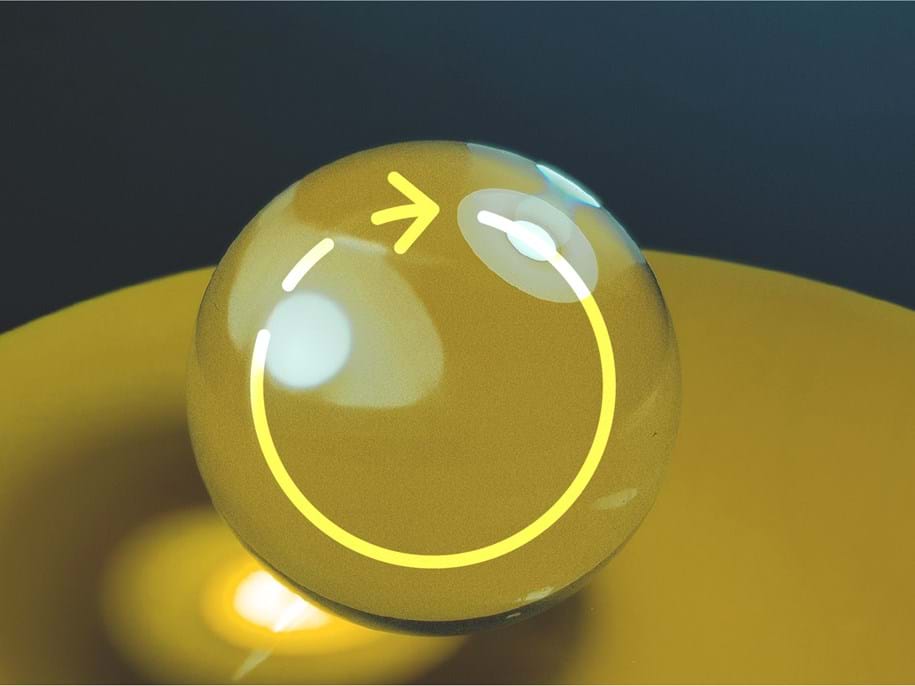 Een reflecterende, transparante bol met een gele pijl en cirkel erop getekend, staat op een geel oppervlak tegen een donkere achtergrond. De pijl wijst omhoog, wat een gevoel van beweging creëert.